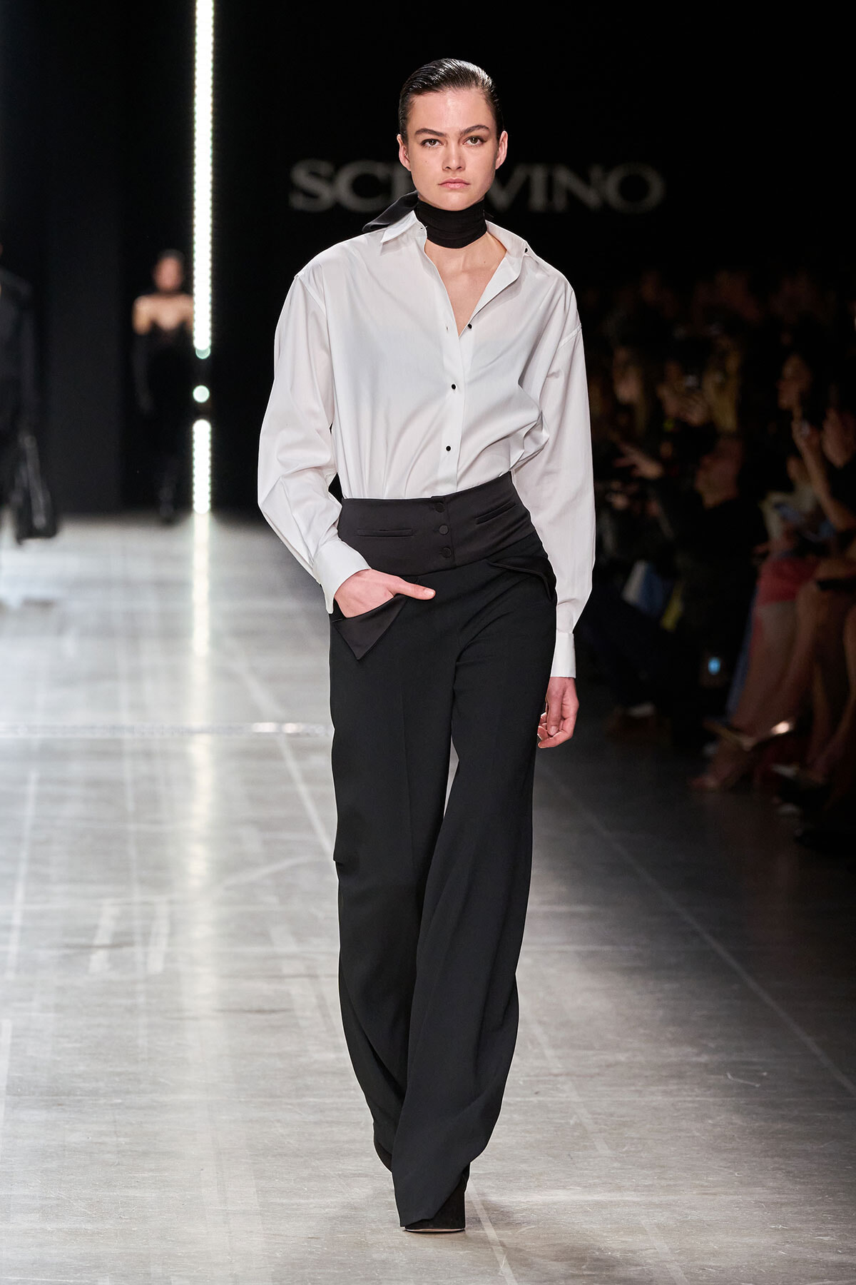 Model walks the runway in a white button-down shirt tucked into high-waisted black wide-leg pants, with a black neck scarf and sleek, pulled-back hair.