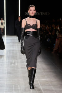 Model on a runway in a dark gothic-inspired outfit: black lace bralette, wide waist belt, long black gloves, gray midi skirt, knee-high boots, choker.