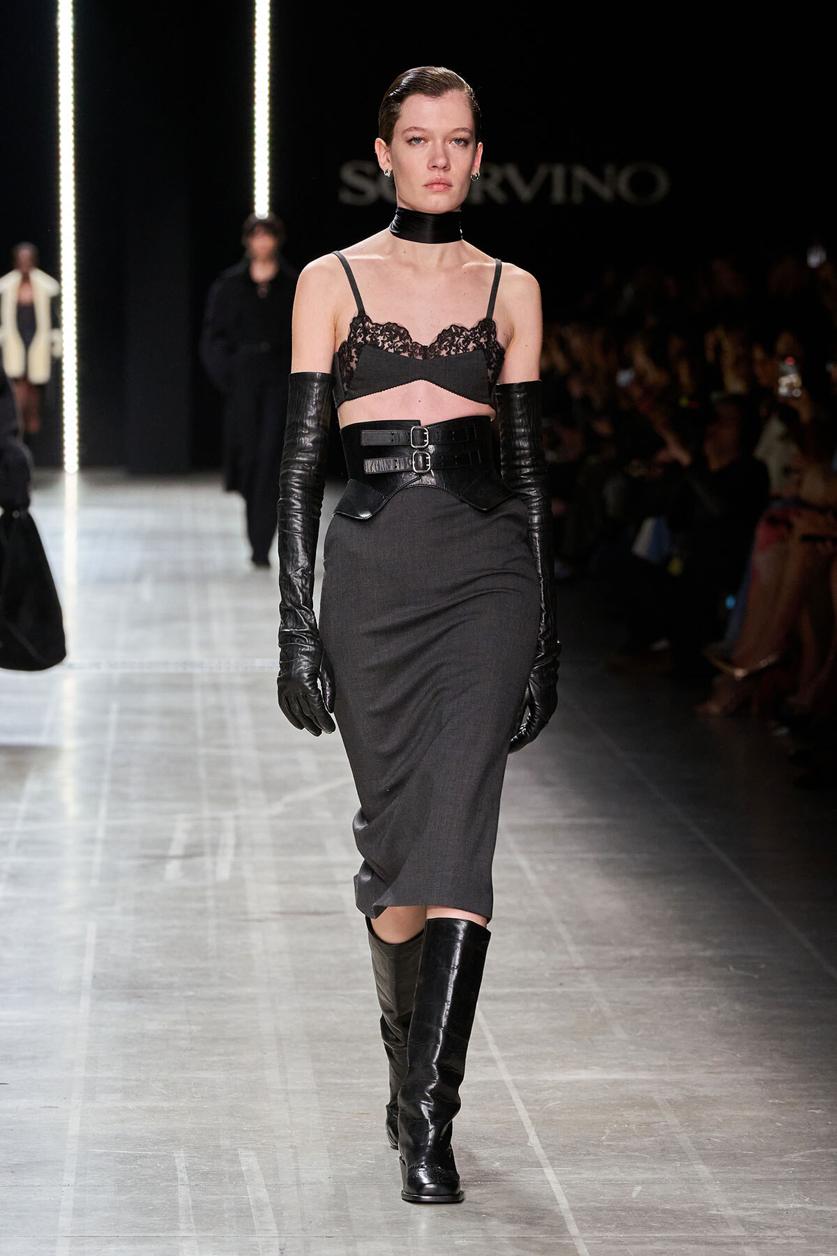 Model on a runway in a dark gothic-inspired outfit: black lace bralette, wide waist belt, long black gloves, gray midi skirt, knee-high boots, choker.