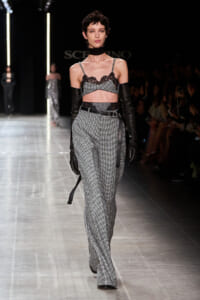 Model walking the runway in a black lace bralette, choker, and high-waisted gray check pants with a belt and long black gloves.
