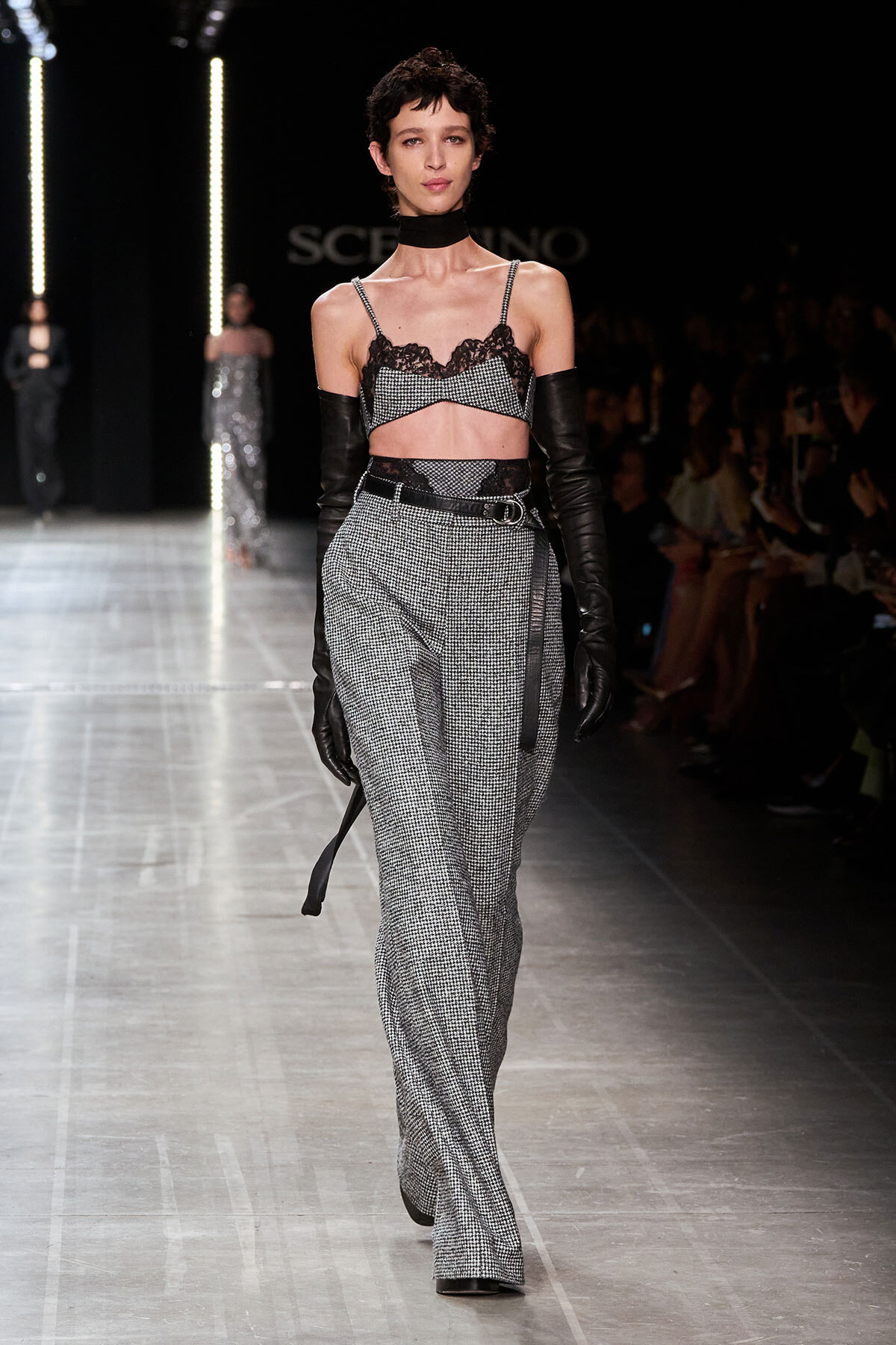 Model walking the runway in a black lace bralette, choker, and high-waisted gray check pants with a belt and long black gloves.