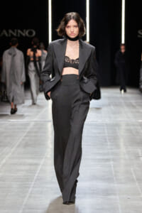 Model walking the runway in a charcoal-gray pantsuit with blazer open to reveal a black lace bralette; hands in pockets.