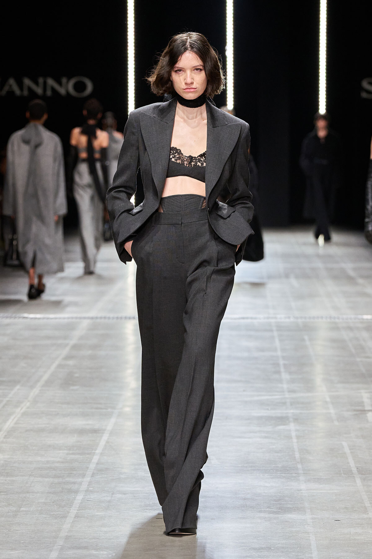 Model walking the runway in a charcoal-gray pantsuit with blazer open to reveal a black lace bralette; hands in pockets.