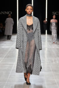 Model on a runway wearing a long gray houndstooth coat over a sheer black lace bodysuit with a choker and black flats.