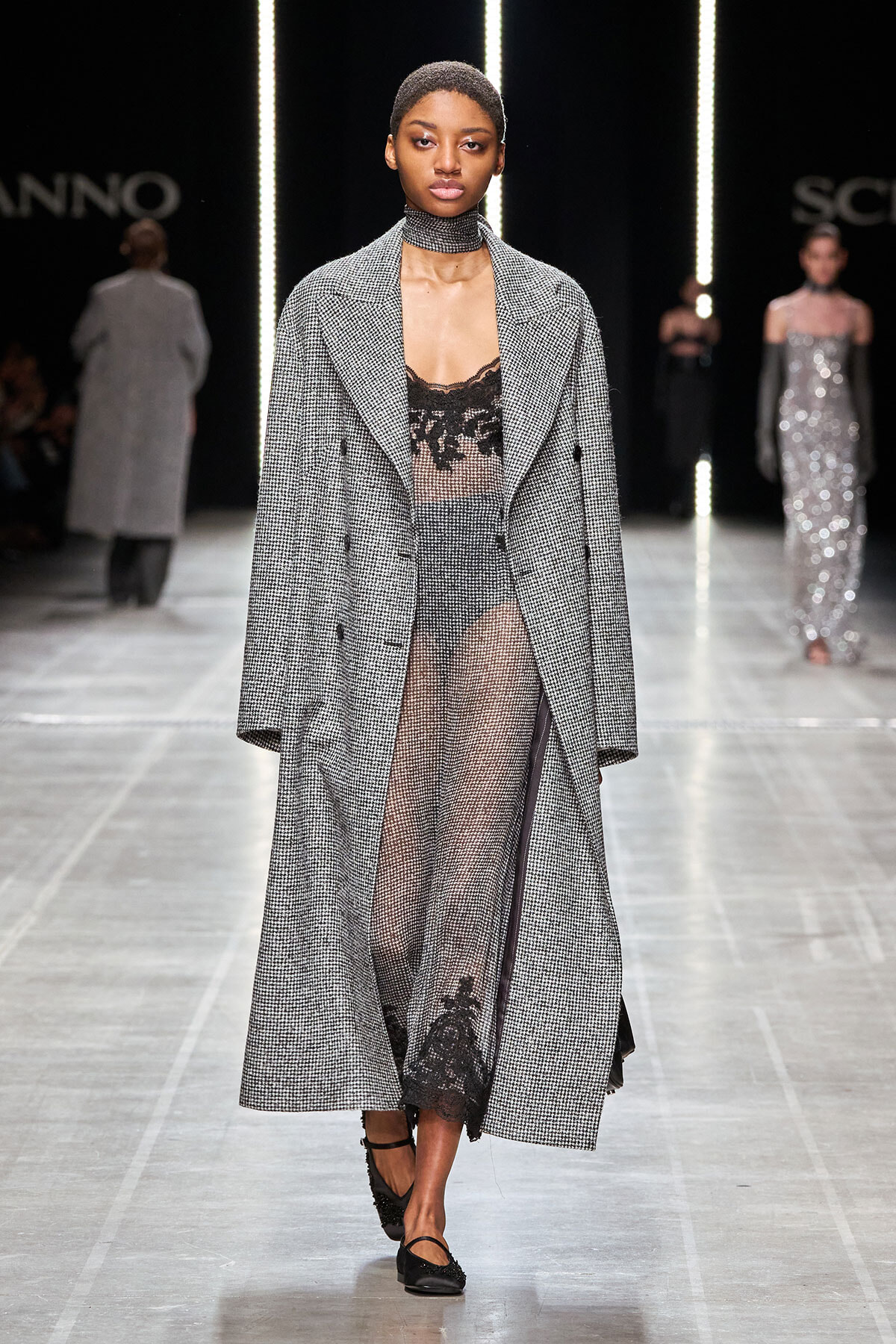 Model on a runway wearing a long gray houndstooth coat over a sheer black lace bodysuit with a choker and black flats.