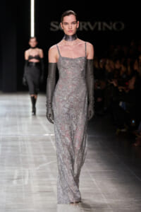 Model walking the runway in a full-length silver sequined gown with thin straps, matching long grey gloves and a metallic choker.