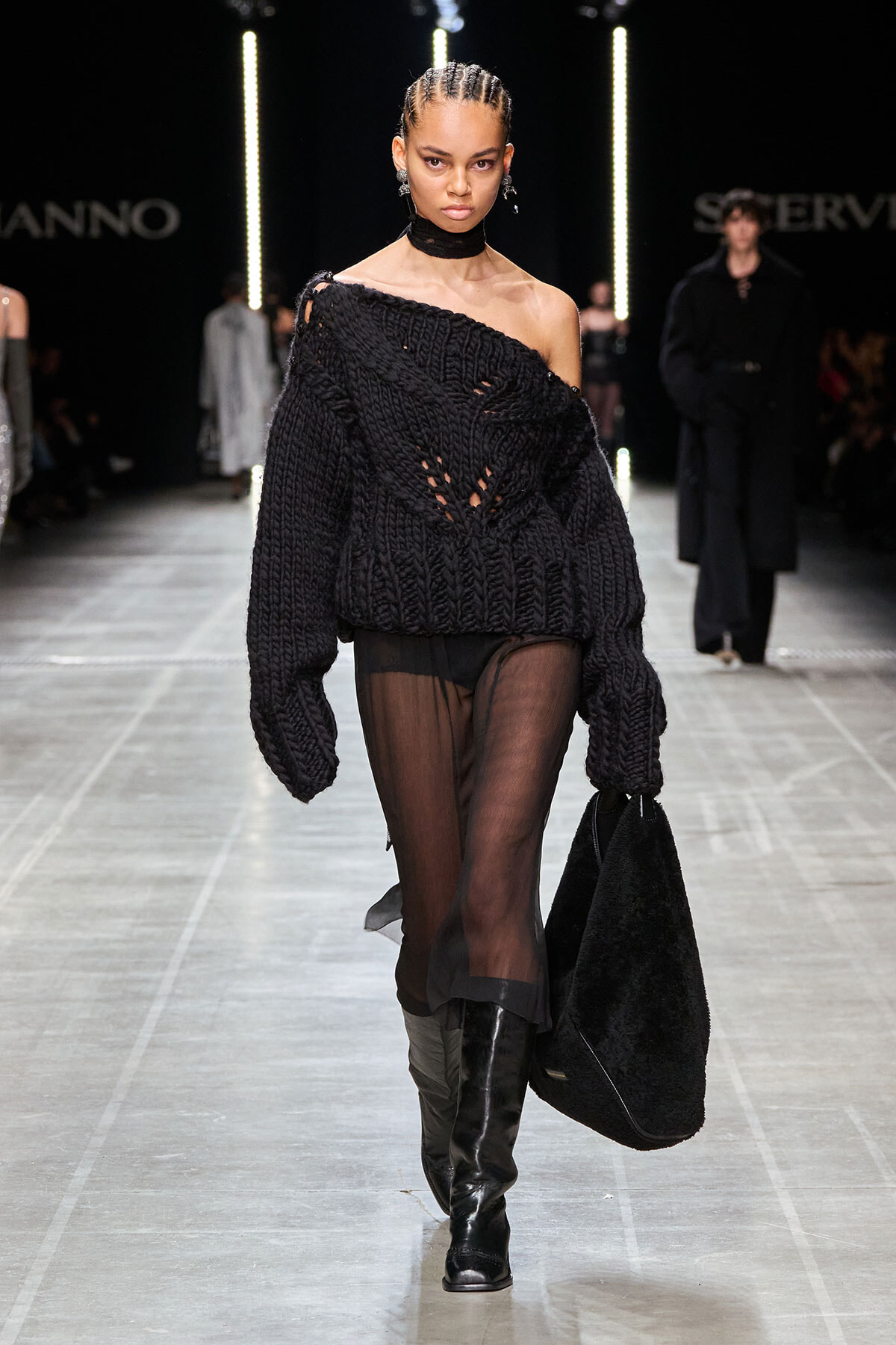 Model on a runway wearing an oversized black knit off-the-shoulder sweater with sheer tights, knee-high boots, and a large black bag.