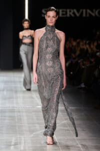 Model walks the runway in a silver, beaded high-neck gown with a sheer, intricate pattern and a long, subtle train.