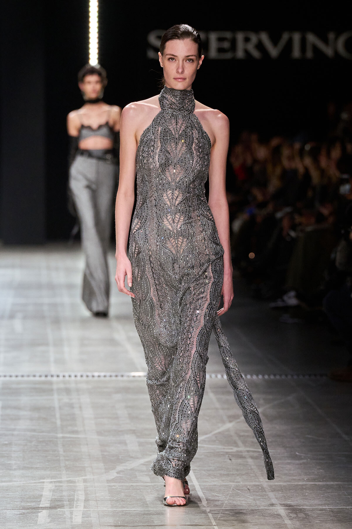 Model walks the runway in a silver, beaded high-neck gown with a sheer, intricate pattern and a long, subtle train.