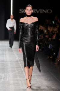Model walks the runway in a black off-shoulder leather dress with a corseted waist and choker, dramatic and sleek.