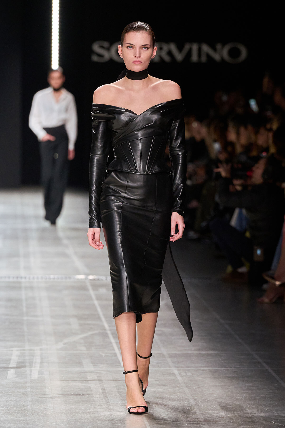 Model walks the runway in a black off-shoulder leather dress with a corseted waist and choker, dramatic and sleek.