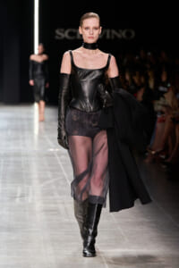 Runway model in a black leather corset, sheer skirt, long gloves, knee-high boots, and a choker on a fashion catwalk.