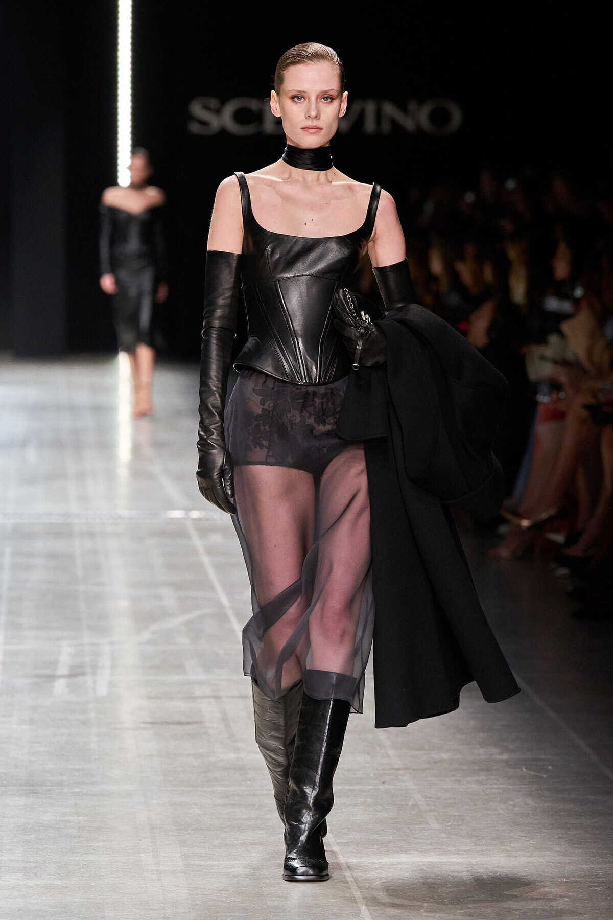 Runway model in a black leather corset, sheer skirt, long gloves, knee-high boots, and a choker on a fashion catwalk.