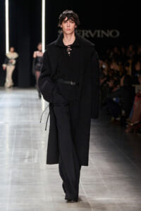 Model walking a monochrome runway in an all-black ensemble: long coat, wide knit sleeves, lace-up top, belted waist.