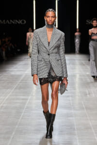 Model walks the runway in a gray plaid blazer worn as a dress over black lace shorts, black boots, and silver jewelry.",