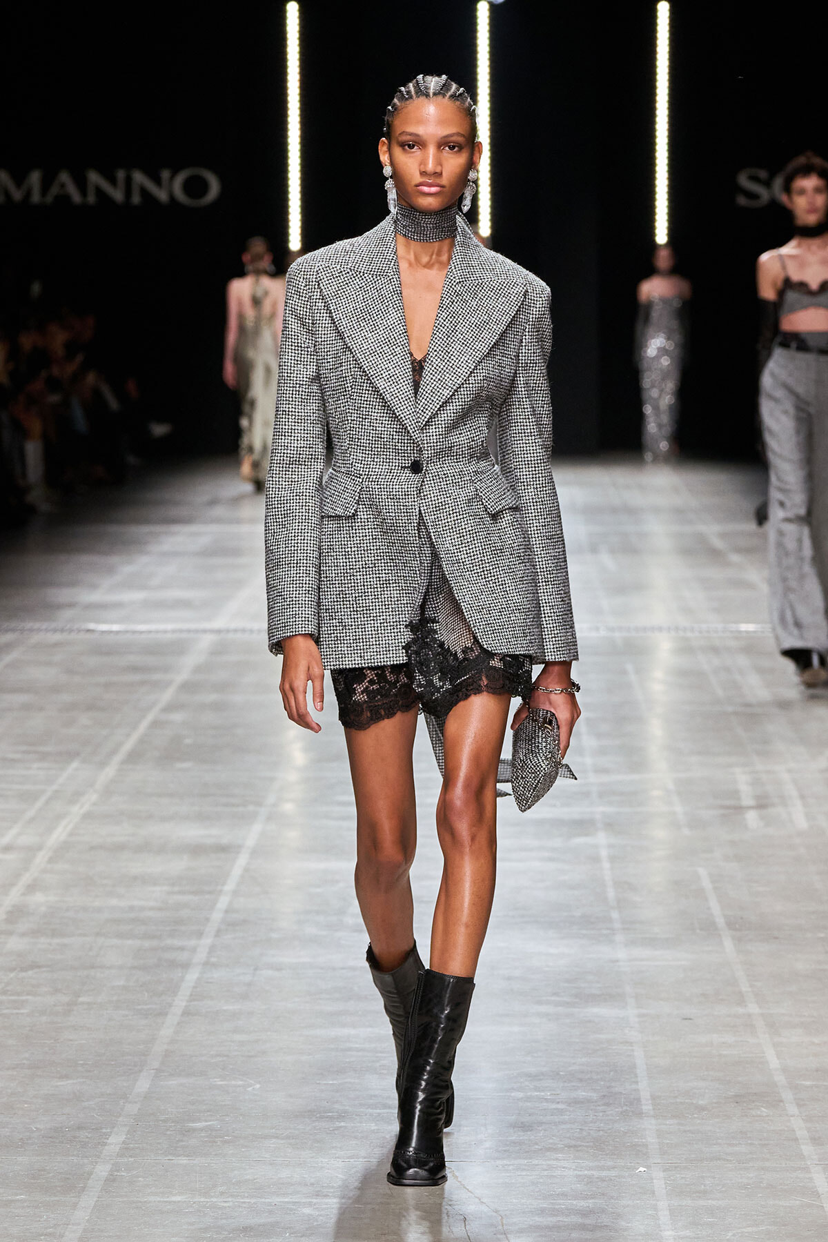 Model walks the runway in a gray plaid blazer worn as a dress over black lace shorts, black boots, and silver jewelry.",