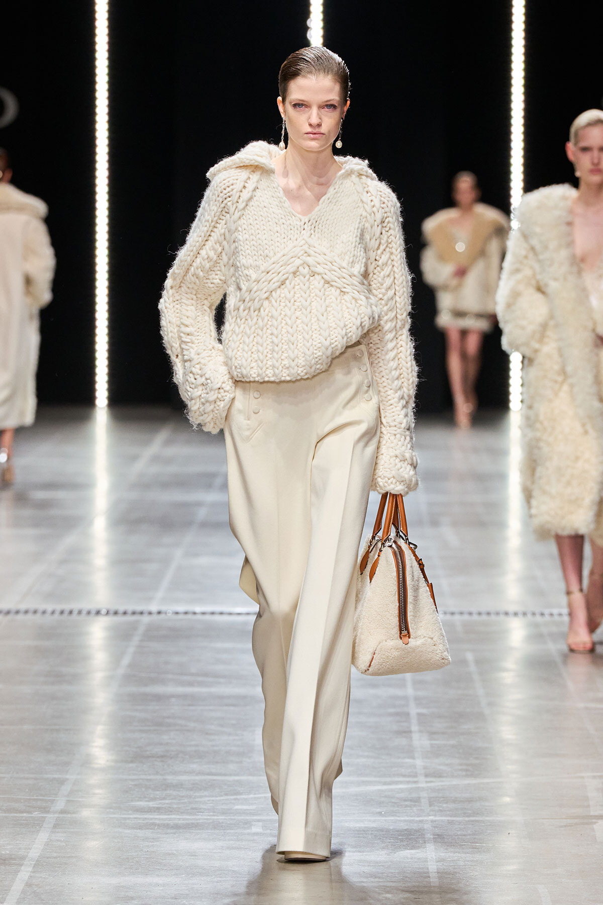 Model walks a runway in a bulky cream knit sweater and wide-leg cream trousers, holding a cream shearling handbag with brown straps.