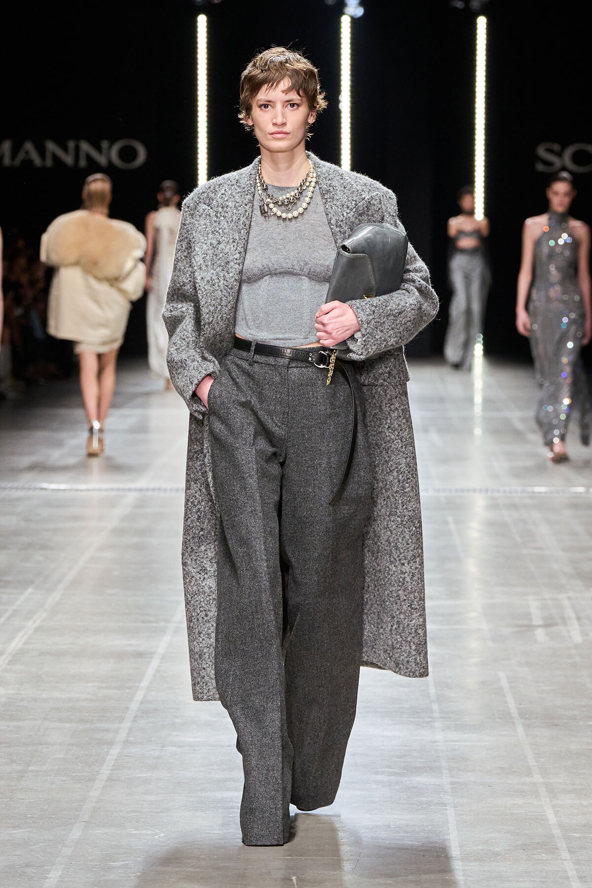 Runway model in a gray textured coat over a matching top and wide-leg trousers, holding a gray clutch and wearing layered pearl necklaces.