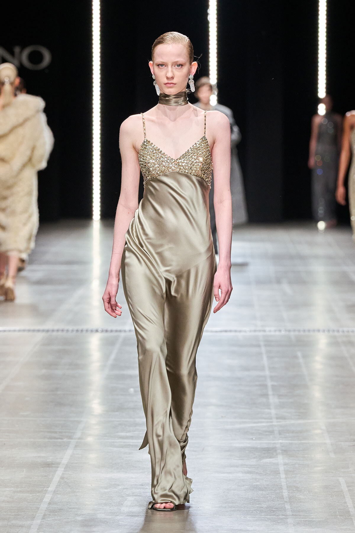Model walks runway in a gold satin gown with a beaded bodice and metallic choker, during a fashion show.