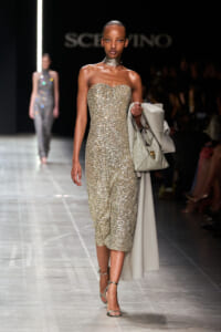 Model walking the runway in a gold sequined strapless midi dress, holding a light gray coat and matching handbag.