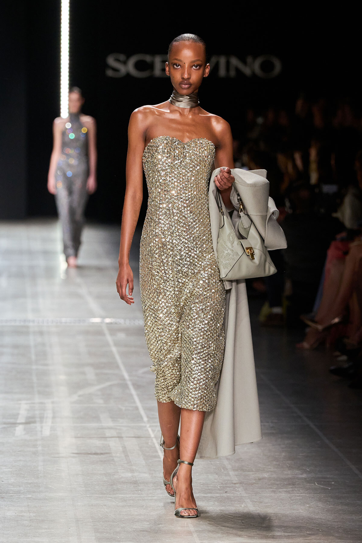 Model walking the runway in a gold sequined strapless midi dress, holding a light gray coat and matching handbag.
