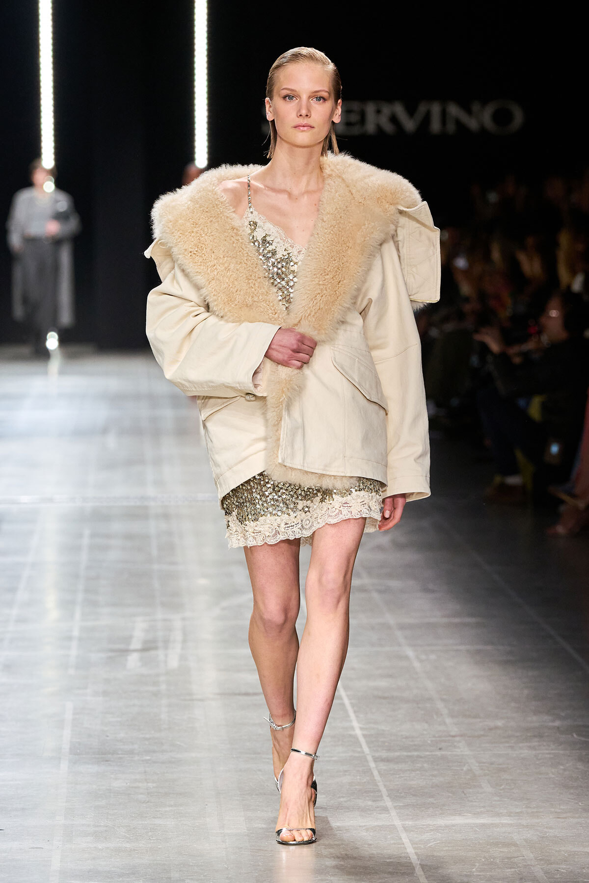 Model walking a fashion runway in a beige oversized coat with a fluffy shearling collar over a sequined slip dress and silver sandals.