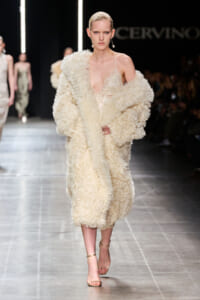 Model walks runway in a cream textured faux-fur coat over a lace slip dress, beige strappy sandals visible.