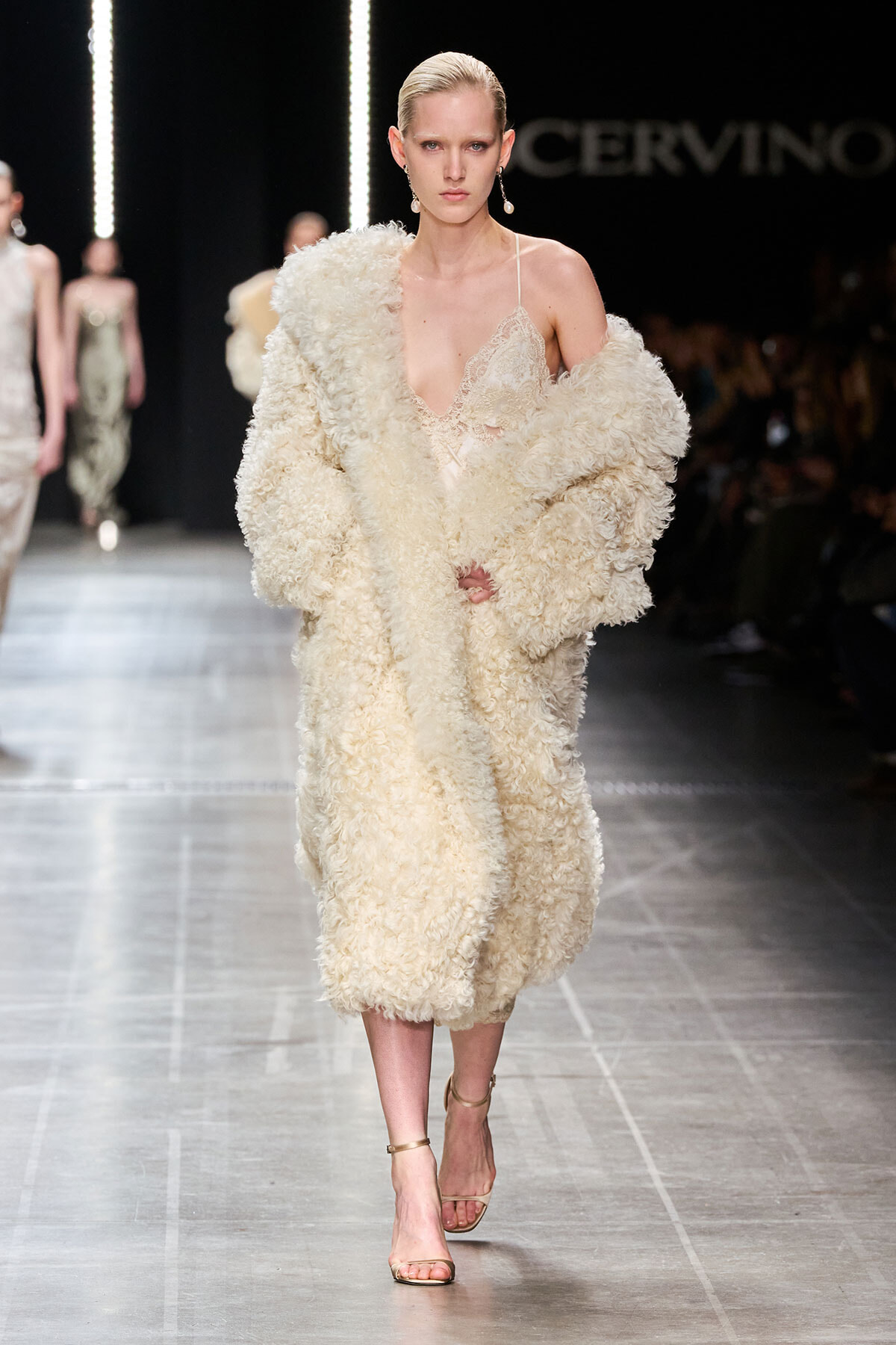 Model walks runway in a cream textured faux-fur coat over a lace slip dress, beige strappy sandals visible.