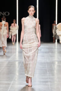 Model walking a runway in a cream lace high-neck sleeveless dress with a wrapped skirt and nude heels, backdrop of other models and vertical light strips.