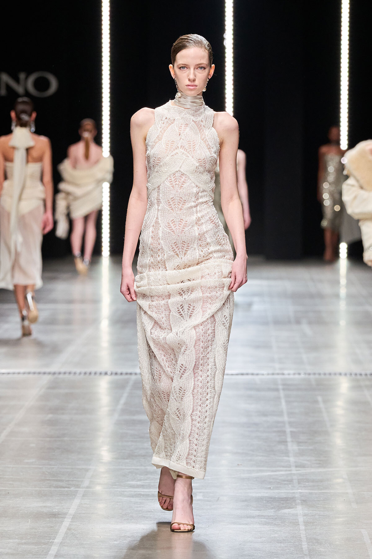 Model walking a runway in a cream lace high-neck sleeveless dress with a wrapped skirt and nude heels, backdrop of other models and vertical light strips.
