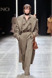 Model walking a runway in a taupe tweed suit with oversized coat, brown belt, and a brown handbag