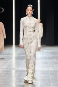 Model walking the runway in an ivory lace-knit, floor-length gown with a high-neck choker and large dangling earrings.