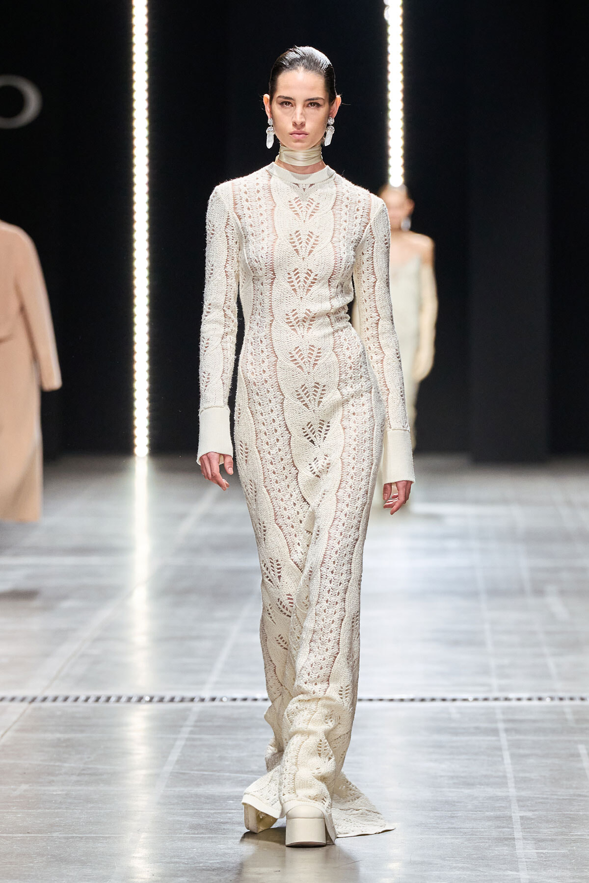 Model walking the runway in an ivory lace-knit, floor-length gown with a high-neck choker and large dangling earrings.