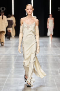 Model on a runway wearing a cream satin slip dress with lace trim, long ivory gloves, and dangling earrings, mid-stride.