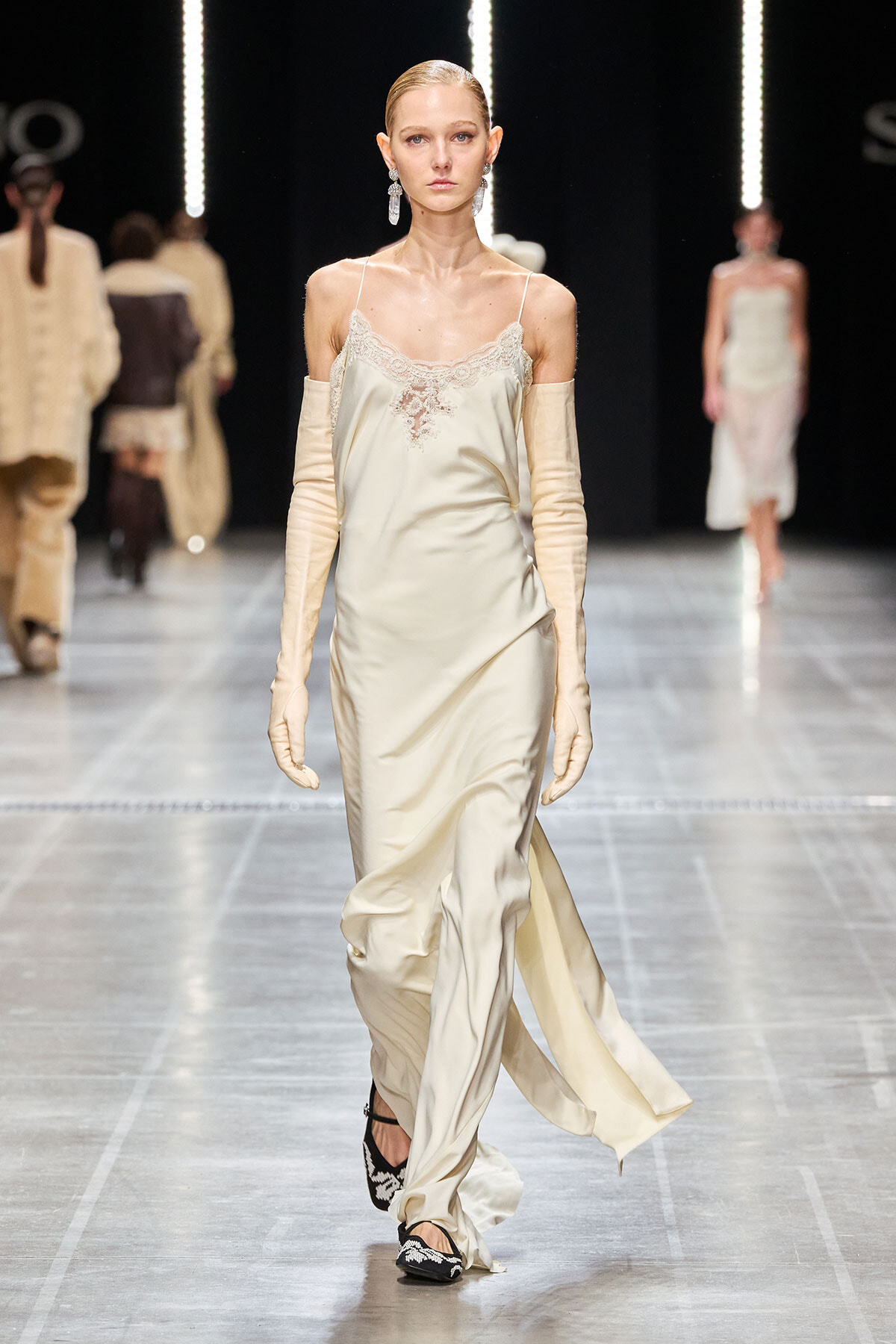 Model on a runway wearing a cream satin slip dress with lace trim, long ivory gloves, and dangling earrings, mid-stride.