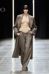 Model walks a fashion runway wearing a taupe oversized coat over a beige corset top and matching wide-leg trousers, hands in pockets, under bright vertical lights.