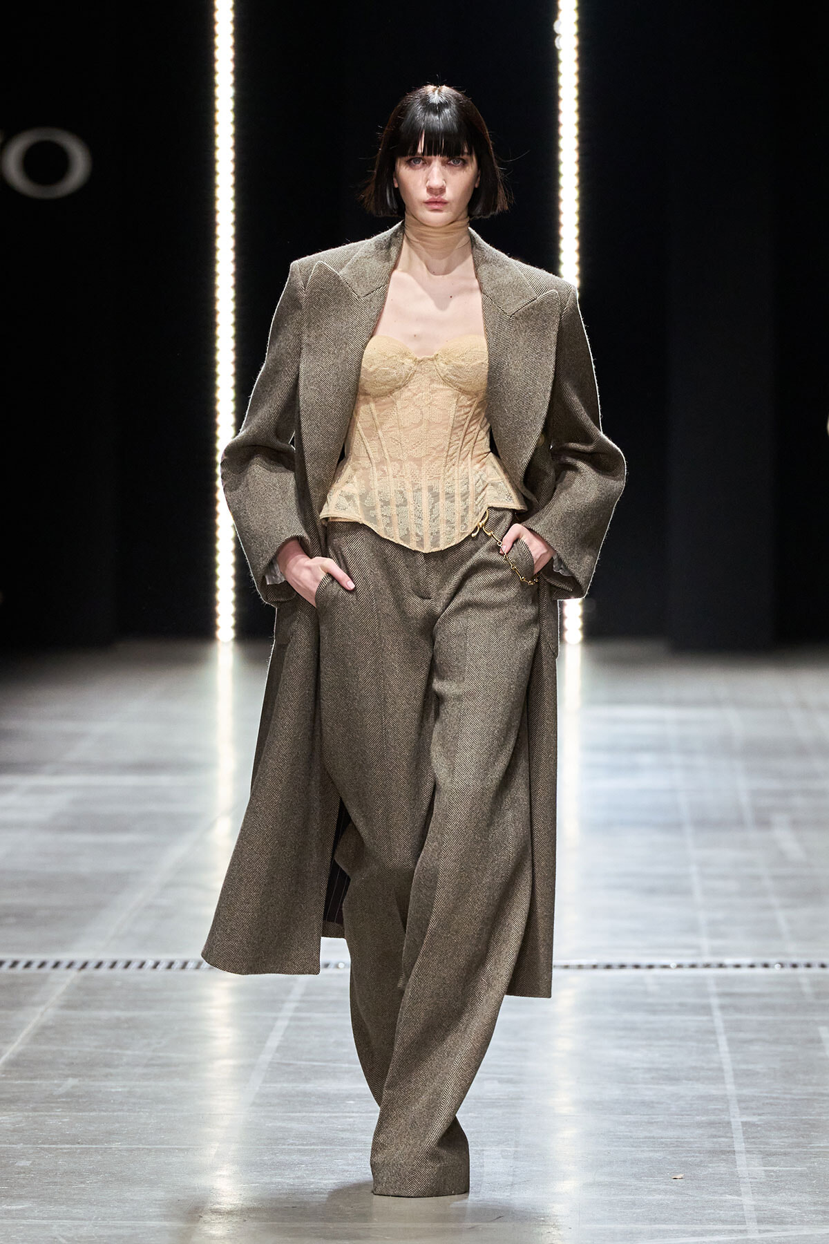 Model walks a fashion runway wearing a taupe oversized coat over a beige corset top and matching wide-leg trousers, hands in pockets, under bright vertical lights.