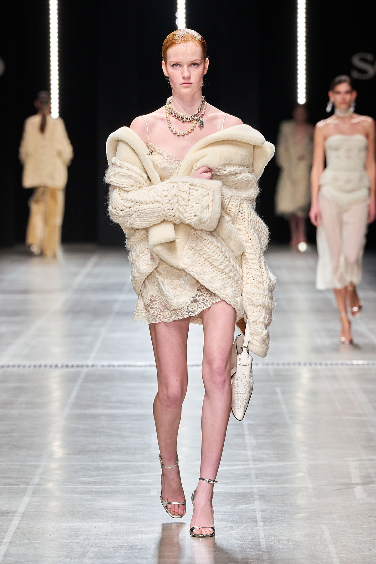Model walks the runway in a voluminous cream knit cardigan draped over an ivory lace dress, accessorized with layered necklaces and silver strappy heels.