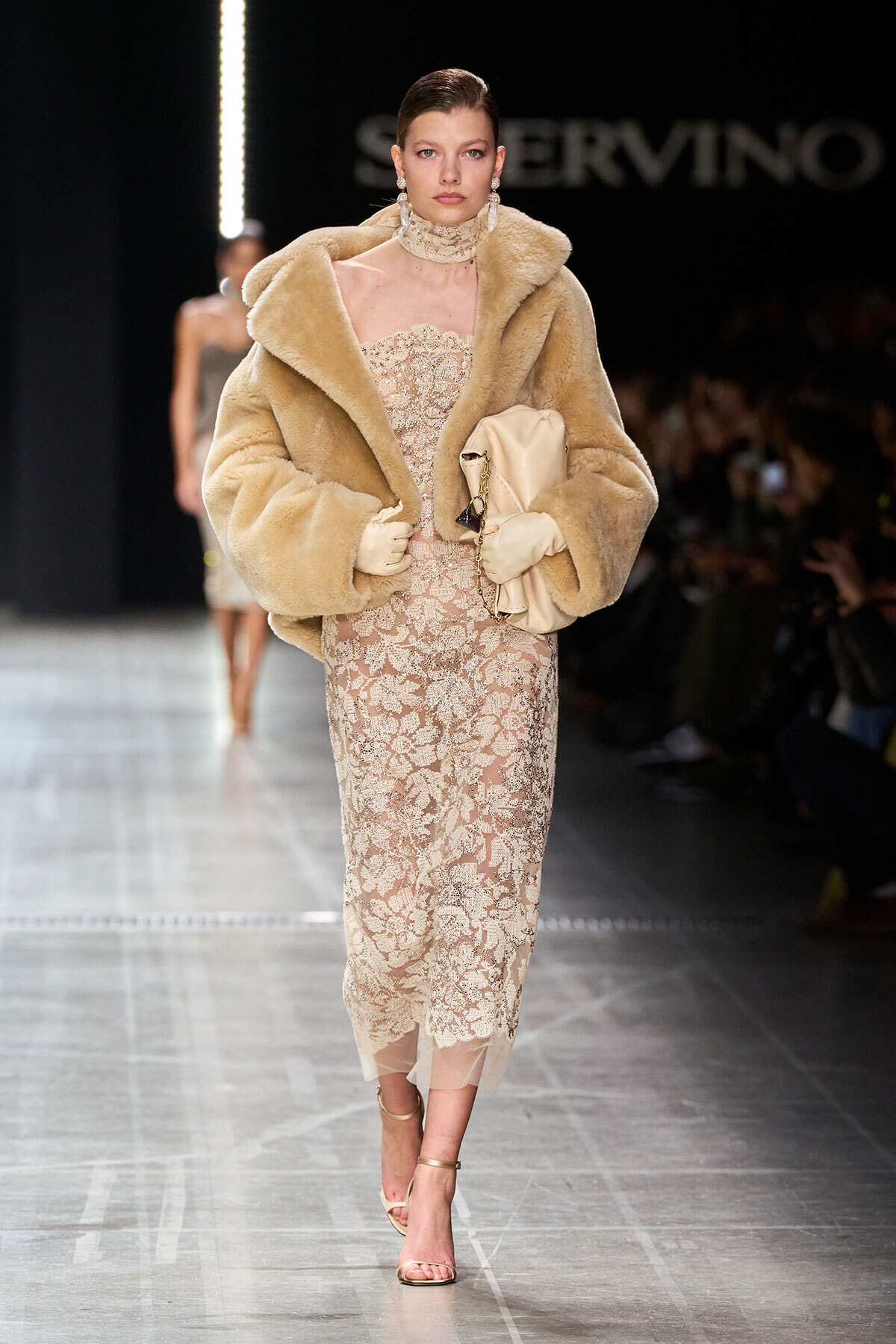 Model walking the runway in a tan fur coat over a champagne lace midi dress, wearing gloves and strappy heels, and carrying a beige clutch.