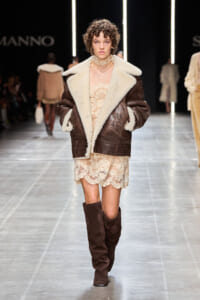 Model on a runway wearing a brown shearling jacket with cream fur lining over a beige lace dress and knee-high brown boots.