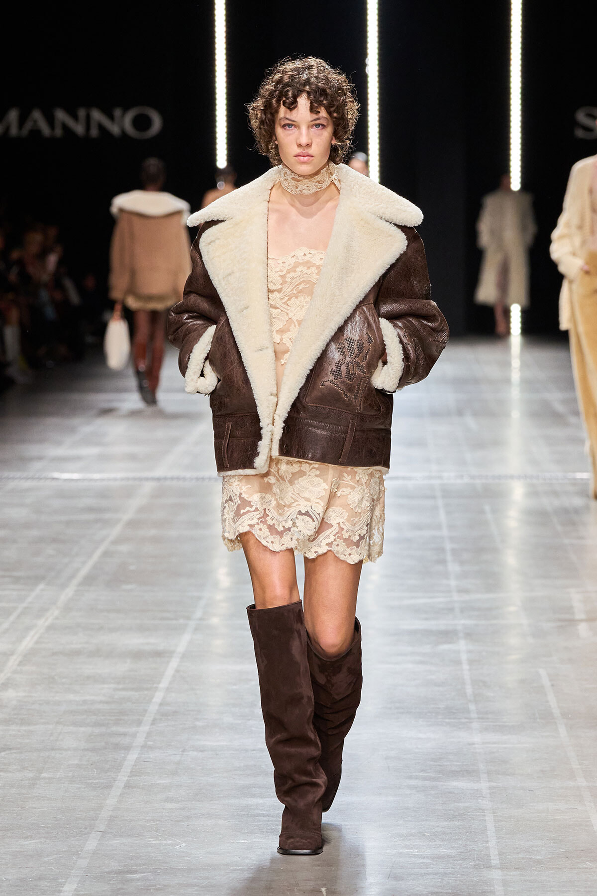 Model on a runway wearing a brown shearling jacket with cream fur lining over a beige lace dress and knee-high brown boots.