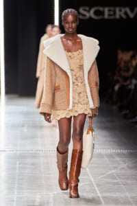 Model walks the runway in camel-toned outfit: a cream shearling‑trimmed coat over a beige lace mini dress, with tall brown boots and a round beige handbag.