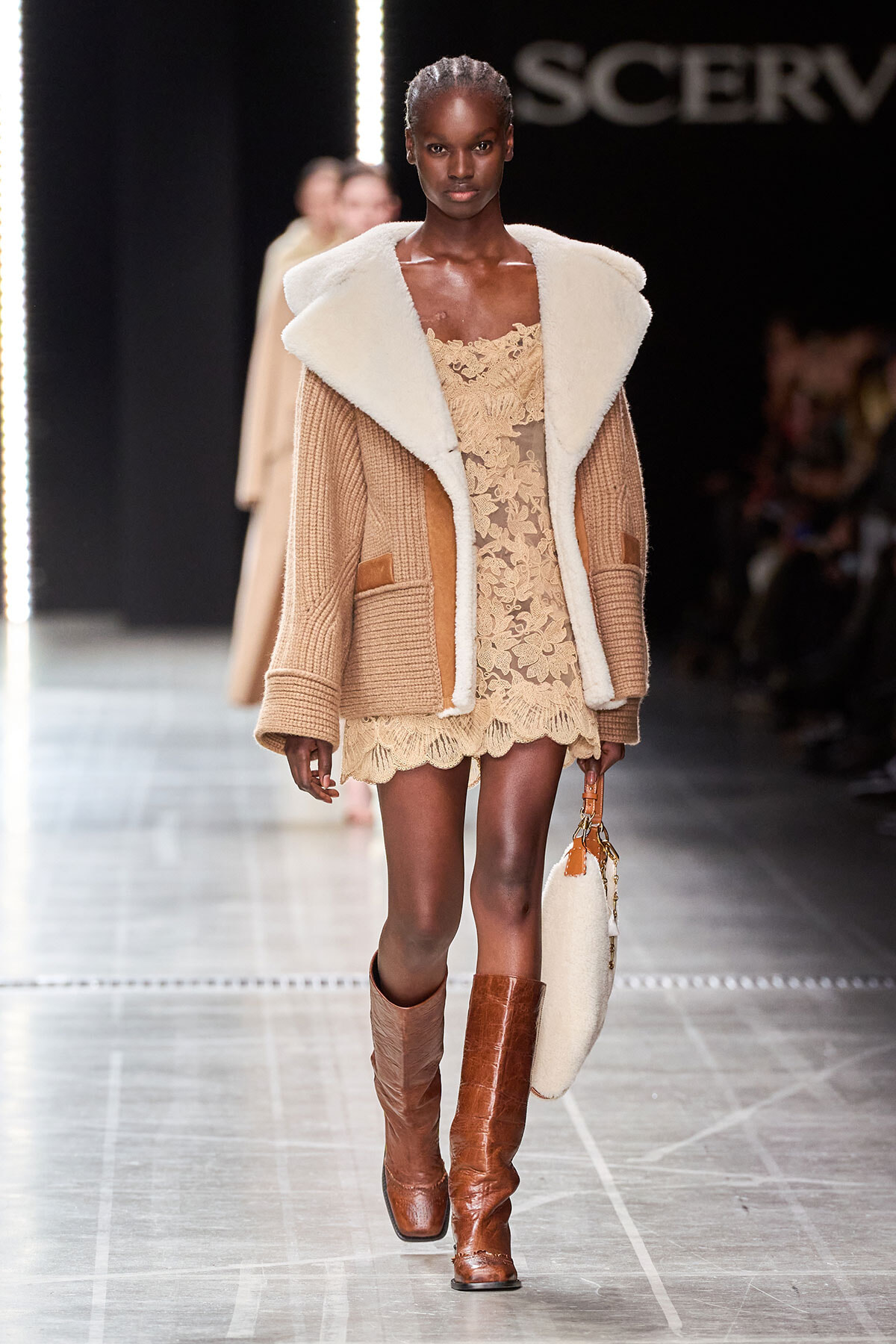 Model walks the runway in camel-toned outfit: a cream shearling‑trimmed coat over a beige lace mini dress, with tall brown boots and a round beige handbag.