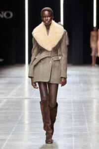 Model on a runway wearing a taupe blazer-style coat with an oversized cream shearling collar and brown knee-high suede boots.