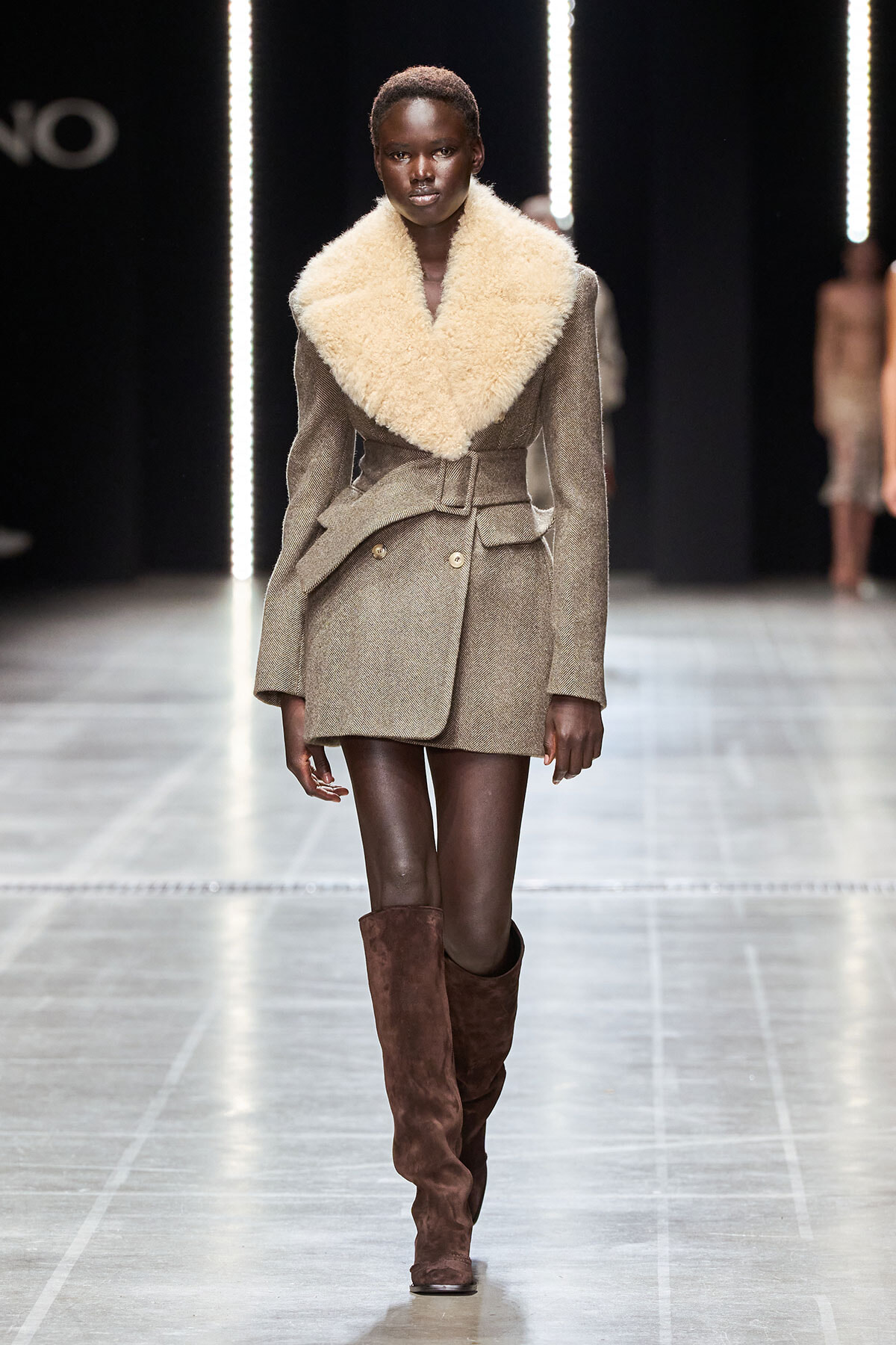 Model on a runway wearing a taupe blazer-style coat with an oversized cream shearling collar and brown knee-high suede boots.