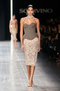 Model on a runway wearing a structured, strapless taupe corset top and a beige lace pencil skirt, paired with a choker and chandelier earrings, walking a glossy catwalk during a fashion show.