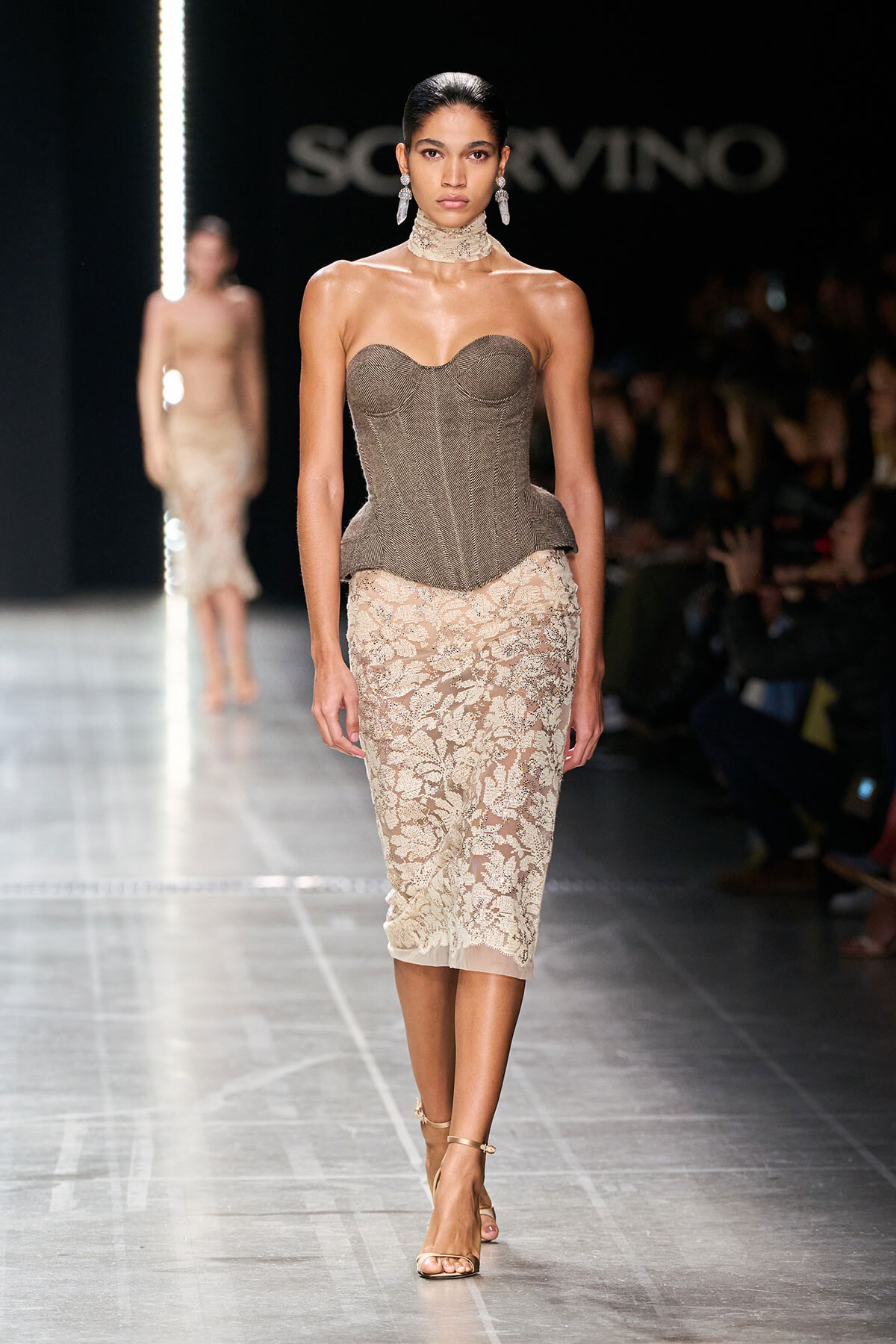 Model on a runway wearing a structured, strapless taupe corset top and a beige lace pencil skirt, paired with a choker and chandelier earrings, walking a glossy catwalk during a fashion show.