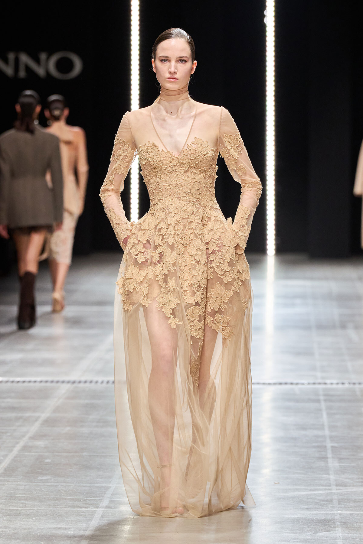 Model walking a nude-toned lace evening gown with sheer overlay on a runway.