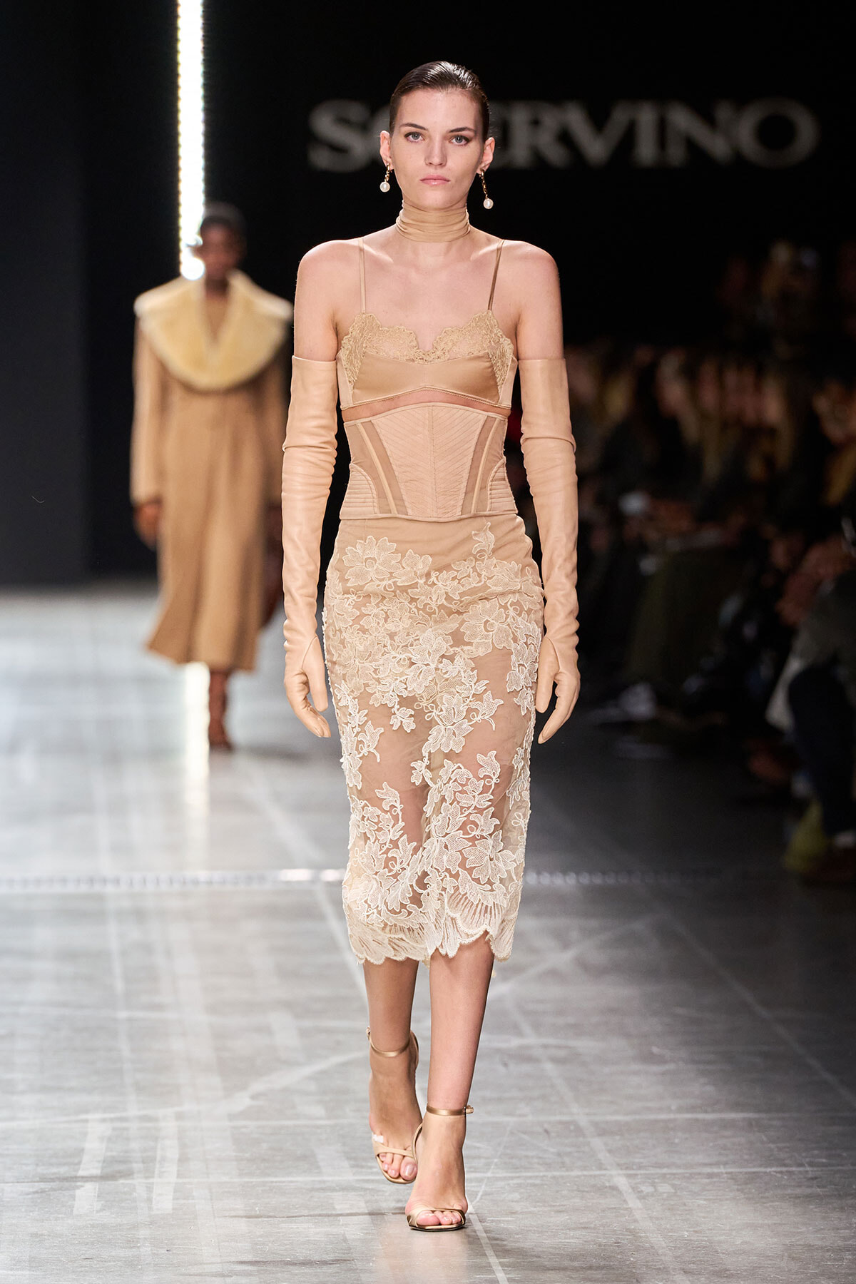 Model walking the runway in a nude-beige lace cocktail dress with sheer panels, long matching gloves, and strappy heels; soft updo and pearl earrings complement the look at a fashion show.