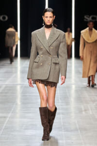 Model walking a runway in a gray tweed double-breasted blazer-dress, with a lace hem peeking below, paired with tall brown suede boots and a dark neck scarf.
